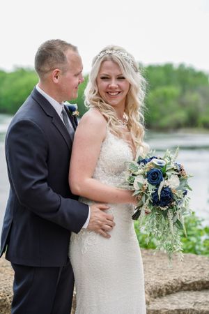 Bride in lace gown holding navy blue bouquet smiles beside groom in dark suit near waterfront.