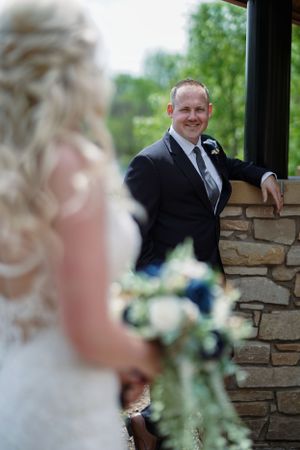 Groom in black suit smiles at blurred bride holding floral bouquet during outdoor wedding ceremony near stone wall.