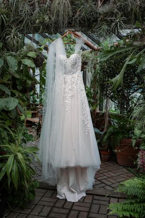 A lace and tulle wedding dress with veil hanging on a wooden hanger in a lush greenhouse setting.
