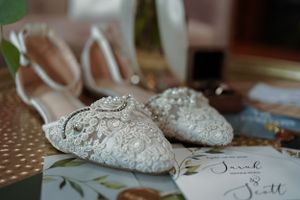 Elegant white lace bridal shoes with pearl embellishments displayed beside wedding stationery with cursive names Sarah and Scott.