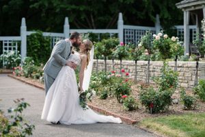 Bride and groom share a romantic kiss in a lush rose garden, with white lattice fencing and blooming flowers.