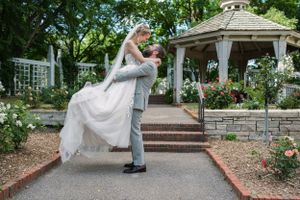 Groom lifts bride in white gown near gazebo surrounded by roses in a romantic outdoor wedding garden setting.