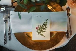 Gold charger plate with white napkin and botanical place card reading Sarah at an elegant wedding table setting.