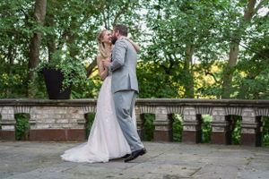 A bride and groom share a romantic dance outdoors on a stone patio surrounded by lush green trees.