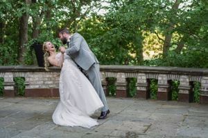 A groom dips his bride for a romantic kiss outdoors, surrounded by lush greenery and stone balustrades.