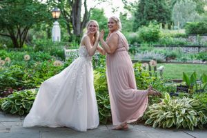 Bride in white gown and bridesmaid in pink dress smiling together in a lush garden setting.