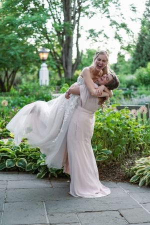 Two brides in elegant gowns joyfully embrace outdoors in a lush garden setting on their wedding day.