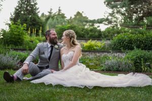 A bride and groom sit together on lush green grass in a garden, sharing a romantic moment in wedding attire.