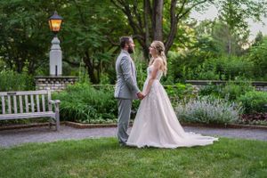 Bride and groom hold hands in a lush garden at dusk, with a glowing lantern and white bench backdrop.
