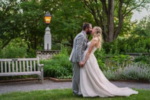 A bride and groom share a romantic kiss in a lush garden setting, with a glowing lantern and white bench nearby.