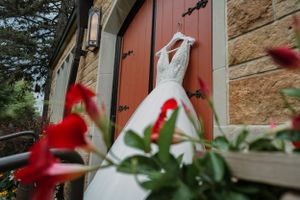 A white wedding dress hanging on a red door of a stone building, framed by vibrant red flowers in the foreground.