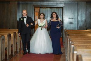 Bride in white ball gown walks down church aisle escorted by parents in formal attire on wedding day.