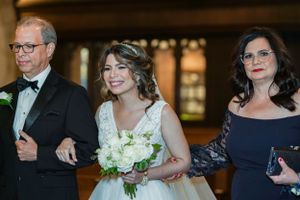 Bride in white lace gown holding bouquet walks down aisle flanked by parents in formal attire at wedding ceremony.