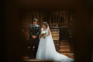 Bride in white gown and groom in dark suit stand together in an ornate wooden church during their wedding ceremony.