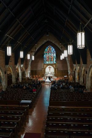Interior of a Gothic church with wooden pews, stained glass window, and a wedding ceremony at the altar.