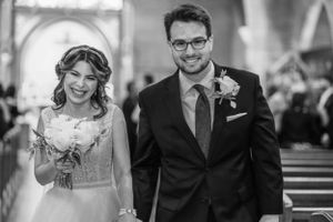Black and white photo of a smiling bride holding a bouquet and groom in suit with boutonniere walking down church aisle.