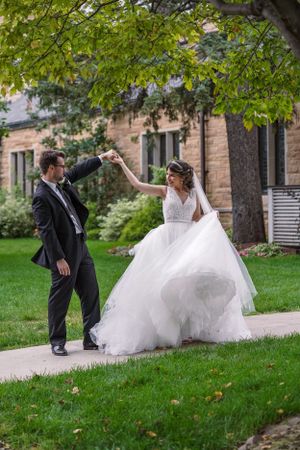 A bride in a flowing white gown twirls with her groom in a black suit on a garden path surrounded by lush greenery.