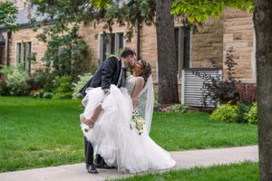 A groom dips his bride for a romantic kiss on a garden pathway outside a stone building on their wedding day.