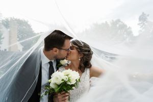 Bride and groom kissing under flowing white veil, bride holding white bouquet on their wedding day.