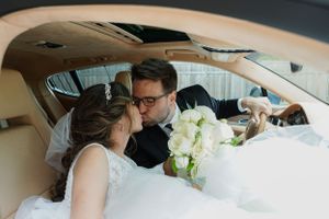 Bride and groom sharing a kiss inside a luxury car, bride holding white rose bouquet on their wedding day.