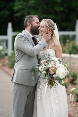 Bearded groom in gray suit tenderly kisses smiling bride holding white and peach floral bouquet in garden.