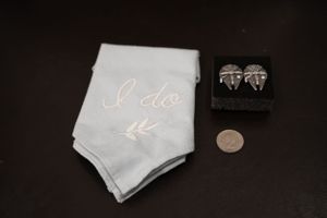 Wedding details flat lay featuring an embroidered I Do handkerchief, silver cufflinks in a box, and a coin on dark background.