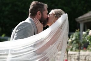 Bride and groom share romantic first kiss as wedding veil billows in the breeze outdoors.