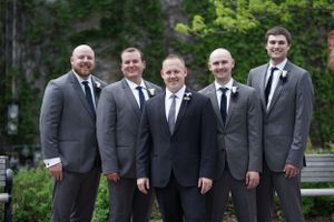 Five groomsmen in matching grey suits with black ties posing outdoors at wedding celebration.