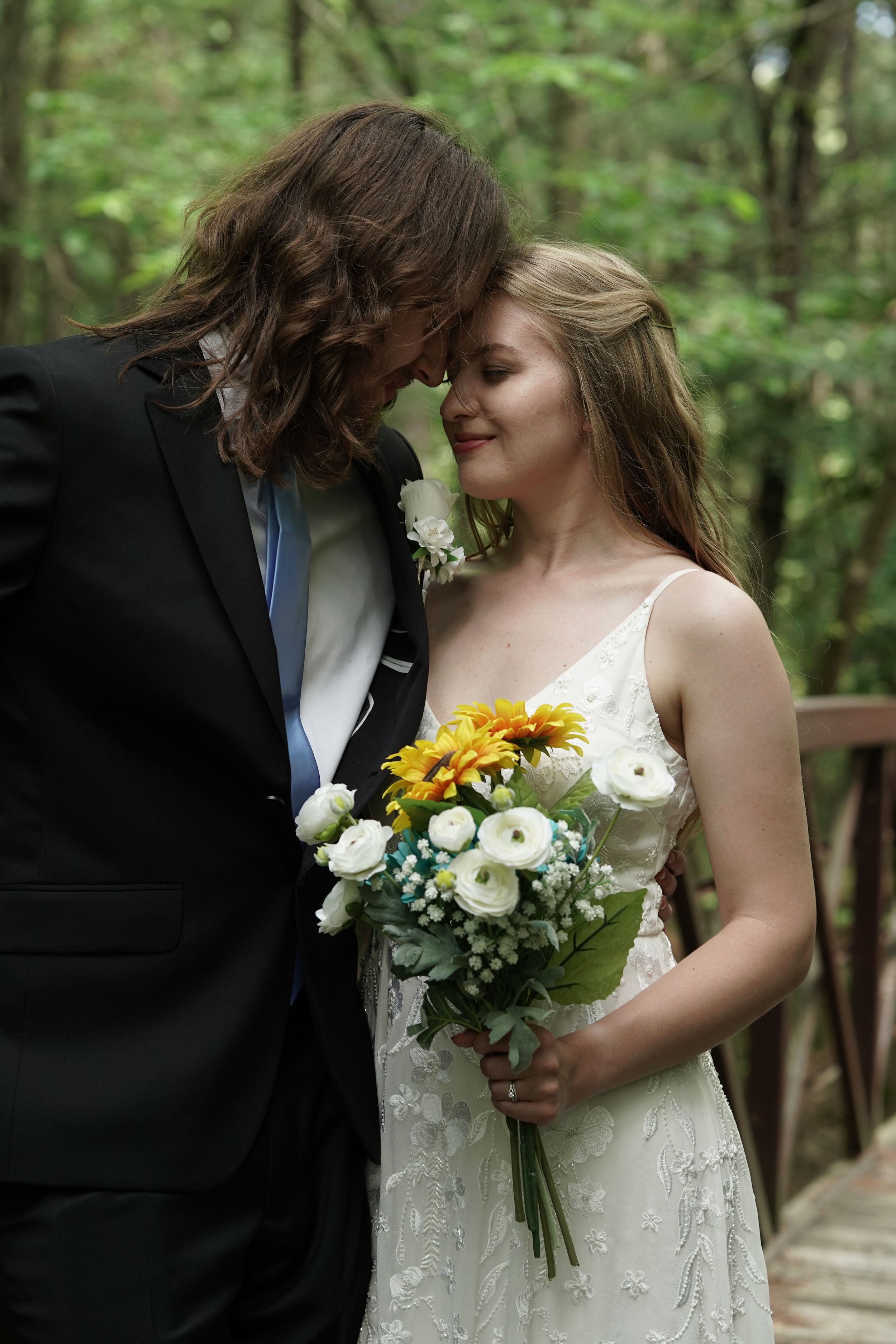Bride and groom touching foreheads tenderly on woodland bridge holding sunflower and white bouquet.