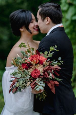 Bride and groom share a romantic kiss while holding a lush red rose and eucalyptus wedding bouquet outdoors.
