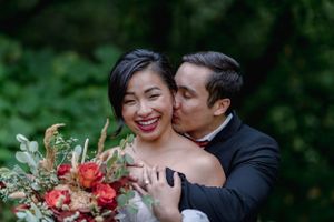 Joyful bride laughs as groom kisses her cheek while she holds a vibrant red and orange bridal bouquet.