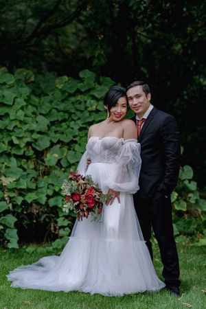 Elegant bride and groom pose together in front of green foliage, bride holding a rich red floral bouquet.
