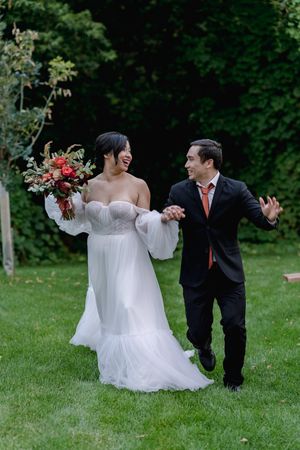 Laughing newlyweds run playfully across a green lawn, bride in a flowing white gown holding a red bouquet.