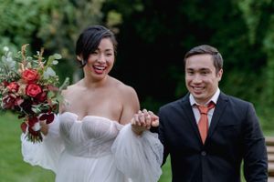 Happy bride and groom walk hand in hand, bride carrying a bold red and green wedding bouquet outdoors.