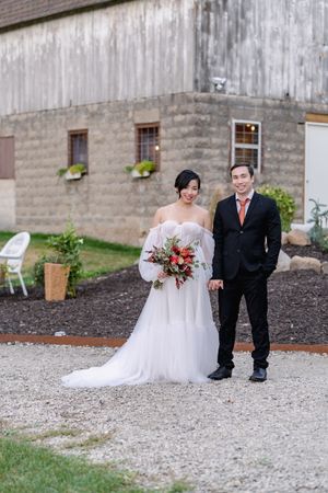 Smiling bride and groom stand together outside a rustic barn, bride holding a stunning red and green bouquet.