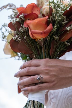 Bride holding autumn bouquet of orange roses with diamond engagement ring visible on hand.