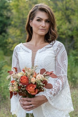 Beautiful bride in boho lace gown holding autumn bouquet of orange roses outdoors.