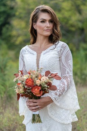 Bride in white lace boho wedding dress holding autumn bouquet of orange roses outdoors.