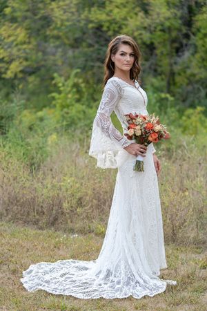 Elegant bride in lace wedding gown with bell sleeves and train holding fall bouquet outdoors.