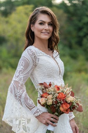Smiling bride in boho lace wedding dress holding orange and rust fall flower bouquet outside.