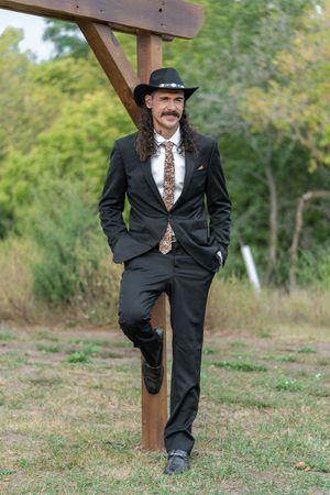 Stylish groom in black suit, cowboy hat, and decorative tie posing by wooden post outside.