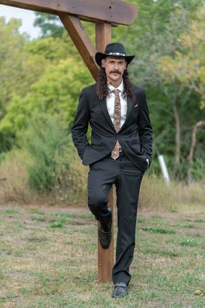 Groom in black suit with cowboy hat and patterned tie leaning on wooden post outdoors.
