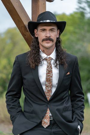 Groom with long hair and mustache in black suit and cowboy hat with floral tie outdoors.