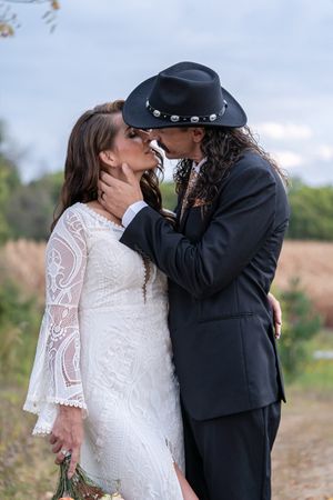Bride and groom sharing a romantic kiss outdoors, groom wearing black cowboy hat and suit, bride in lace wedding dress.