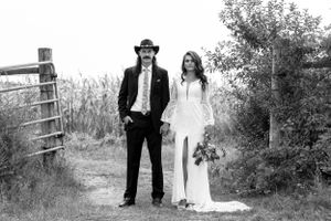 Black and white photo of bride and groom standing on a rural path near a wooden fence and cornfield.