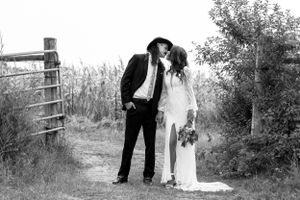 Black and white photo of bride and groom sharing a kiss near a wooden fence with cornfield and trees nearby.