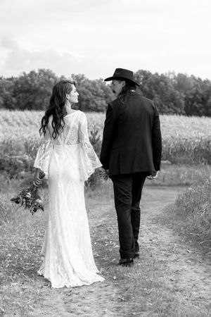 Black and white photo of bride and groom walking hand in hand along a path beside a golden cornfield.