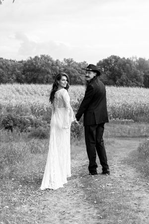 Black and white wedding photo of bride looking back while holding groom's hand on a rural path by a cornfield.