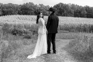 Black and white wedding photo of a bride and groom standing on a path beside a golden wheat field.