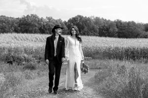 Black and white photo of a smiling bride and groom walking hand in hand along a field path.